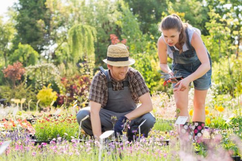 A man and a woman are working together in a well-maintained garden, surrounded by a variety of flowering plants and green foliage. The man, wearing a straw hat, a checked shirt, and gardening gloves, is kneeling on the grass, tending to flower beds that contain a mix of colourful blooms, including pinks, purples, and yellows. The woman, dressed in a denim dress and gardening gloves, is standing and leaning forward, holding garden shears as she trims or shapes a plant. The garden features a lush green lawn area in the foreground, with a backdrop of trees, shrubbery, and a neat hedge line, indicating a landscaped outdoor space typical of residential gardens in Tottenham. Bright sunlight illuminates the scene, creating a vibrant and inviting atmosphere indicative of spring or summer weather. This outdoor environment reflects professional gardening practices, as might be carried out by Lawn Mowing Tottenham, focusing on plant care, lawn maintenance, and garden aesthetics, with natural colour variations and detailed plant textures complementing the tidy, landscaped yard.