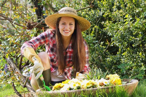 A young woman with long brown hair, smiling and wearing a wide-brimmed straw hat, a red and white checked shirt, and gardening gloves, is crouched down in a lush, well-maintained backyard garden. She is tending to a flower bed filled with bright yellow and white flowers, which are surrounded by a wooden border. The garden features a dense hedge and various small trees or shrubs in the background, with vibrant green foliage. The soil appears healthy, and the grass lawn in the foreground is evenly cut. Natural sunlight illuminates the scene, highlighting the lush greenery and colourful blooms, reflecting a peaceful outdoor gardening environment typical of residential gardens in Tottenham or the surrounding area. This image is relevant to gardening and lawn care services provided by Lawn Mowing Tottenham.