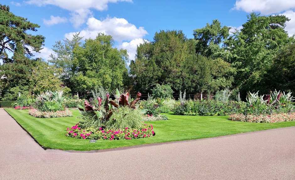 A neatly maintained garden features lush green grass bordered by vibrant flower beds filled with a variety of colorful flowers, including pink and purple blooms. The garden showcases well-trimmed hedg